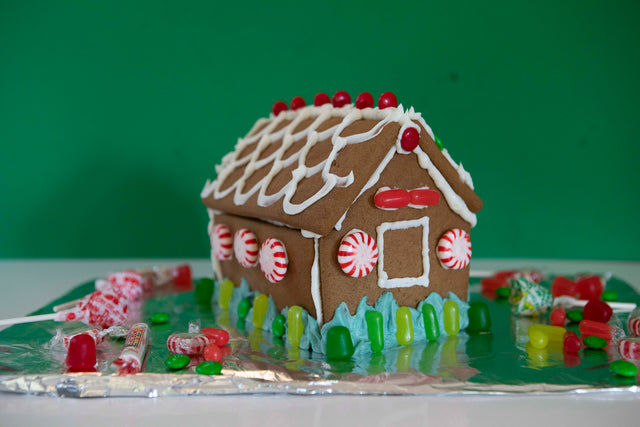 Decorated gingerbread house on top of tin foil with a green backdrop. It is decorated with white and blue icing, peppermints, and green and red candies.