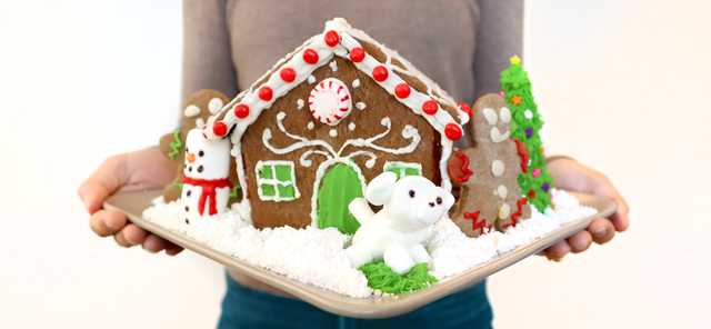 A woman holding up a decorated gingerbread house with winter-themed decorations.